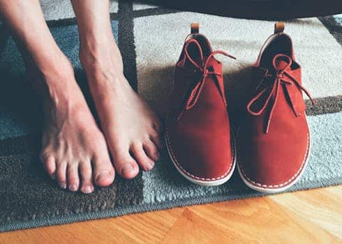 Close-up of bare feet next to red leather shoes on a patterned rug indoors.